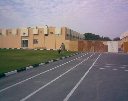 Man walking in a parking lot next to a beige apartment complex with a grassy lawn and a cloudy sky.