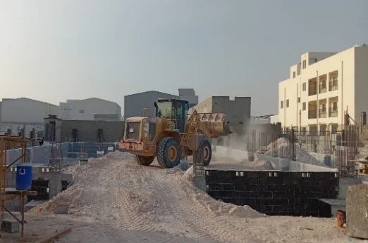 A construction site with a bulldozer pushing dirt and dust, surrounded by buildings and construction materials.