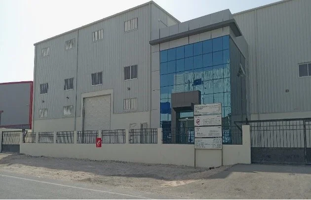 A large industrial building with gray metal siding and a main entrance with reflective blue glass panels, a signboard outside, and a black metal fence in front.