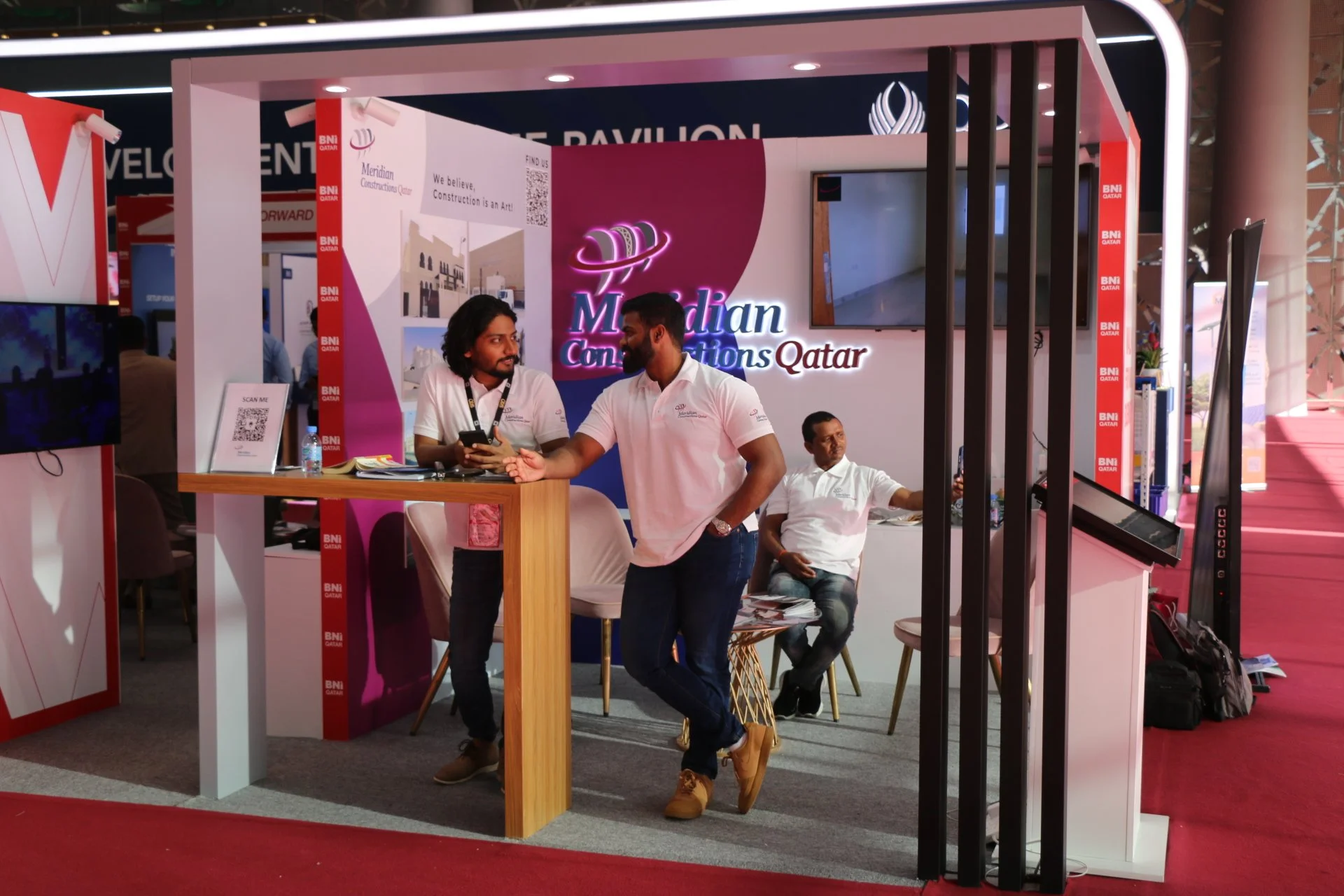 Booth at a trade show with the logo 'Meridian Constructions Qatar' and three men in white shirts, one sitting and two standing, engaged in conversation.