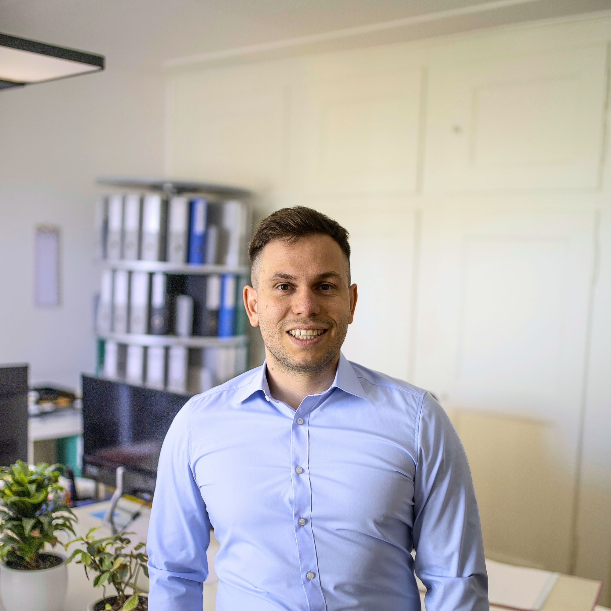 Ein junger Mann trägt ein hellblaues Hemd und steht in einem Büro vor einer weißen Wand mit Bilderrahmen. Im Hintergrund sind Aktenordner, Schreibtisch und Pflanzen sichtbar.