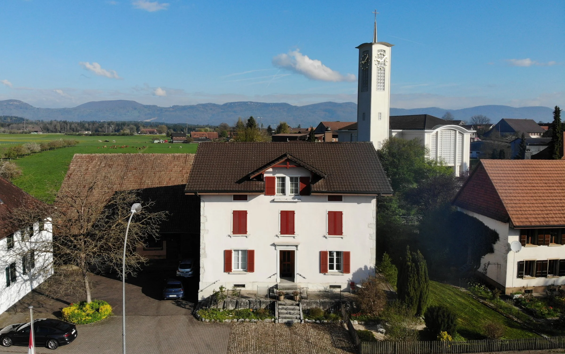 Eine weiße Kirche mit hohen spitzen Türmen und einer Uhr, umgeben von Häusern in einem ländlichen Gebiet, mit Bergen im Hintergrund, bei sonnigem Himmel.