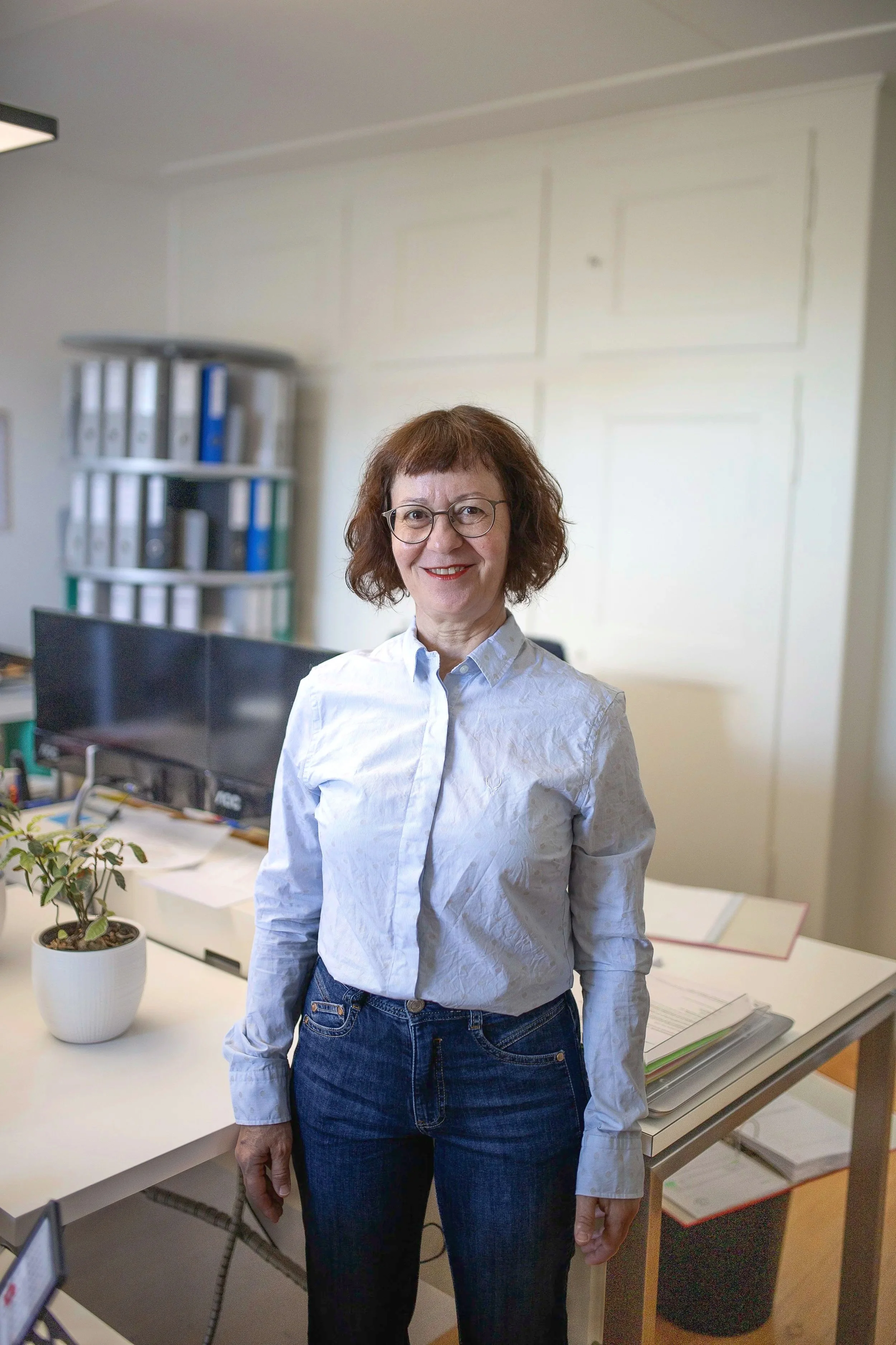 Eine Frau mit Brille und braunen, lockigen Haaren steht in einem Büro vor einem Schreibtisch. Sie trägt eine weiße Hemdbluse und dunkle Jeans. Im Hintergrund sind Aktenordner und ein Computermonitor zu sehen.