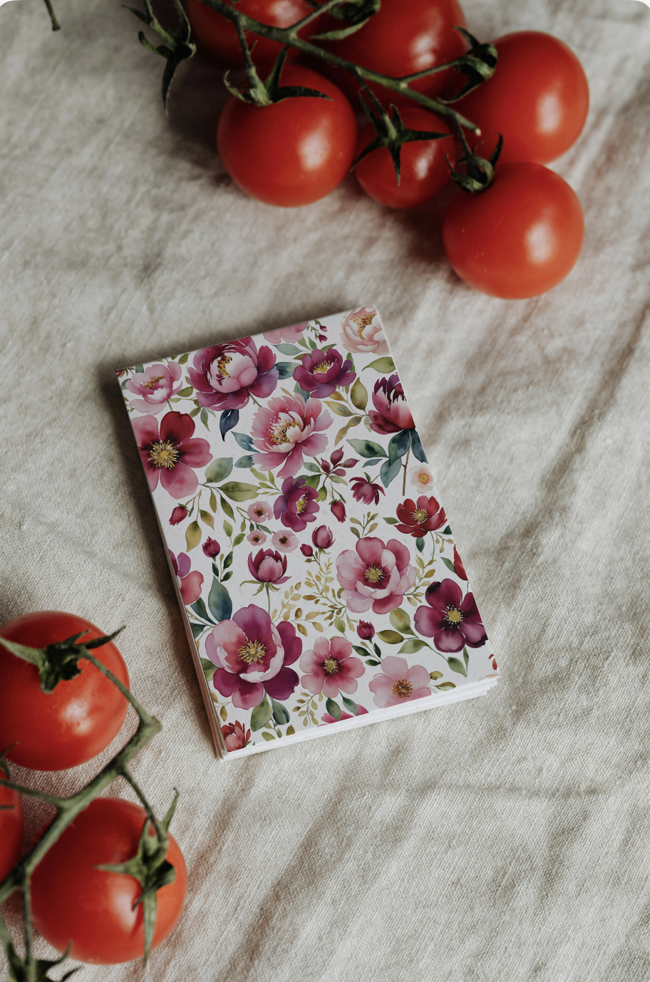 A notebook with a floral cover design, surrounded by tomatoes on a wooden surface.