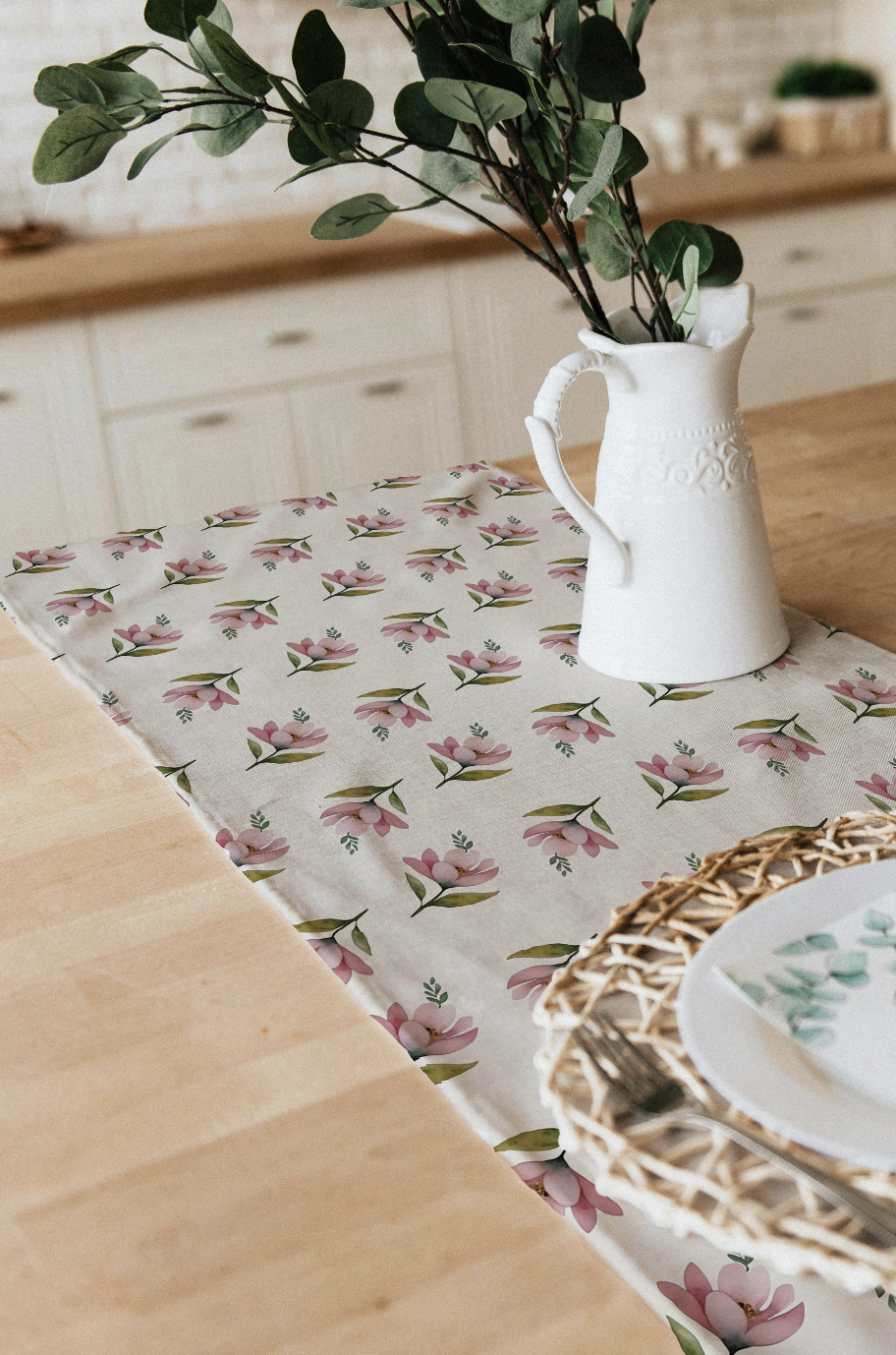 A floral table runner with pink flowers and green leaves on a wooden table, with a white pitcher holding green leafy branches, a woven placemat, and a plate with a fork.