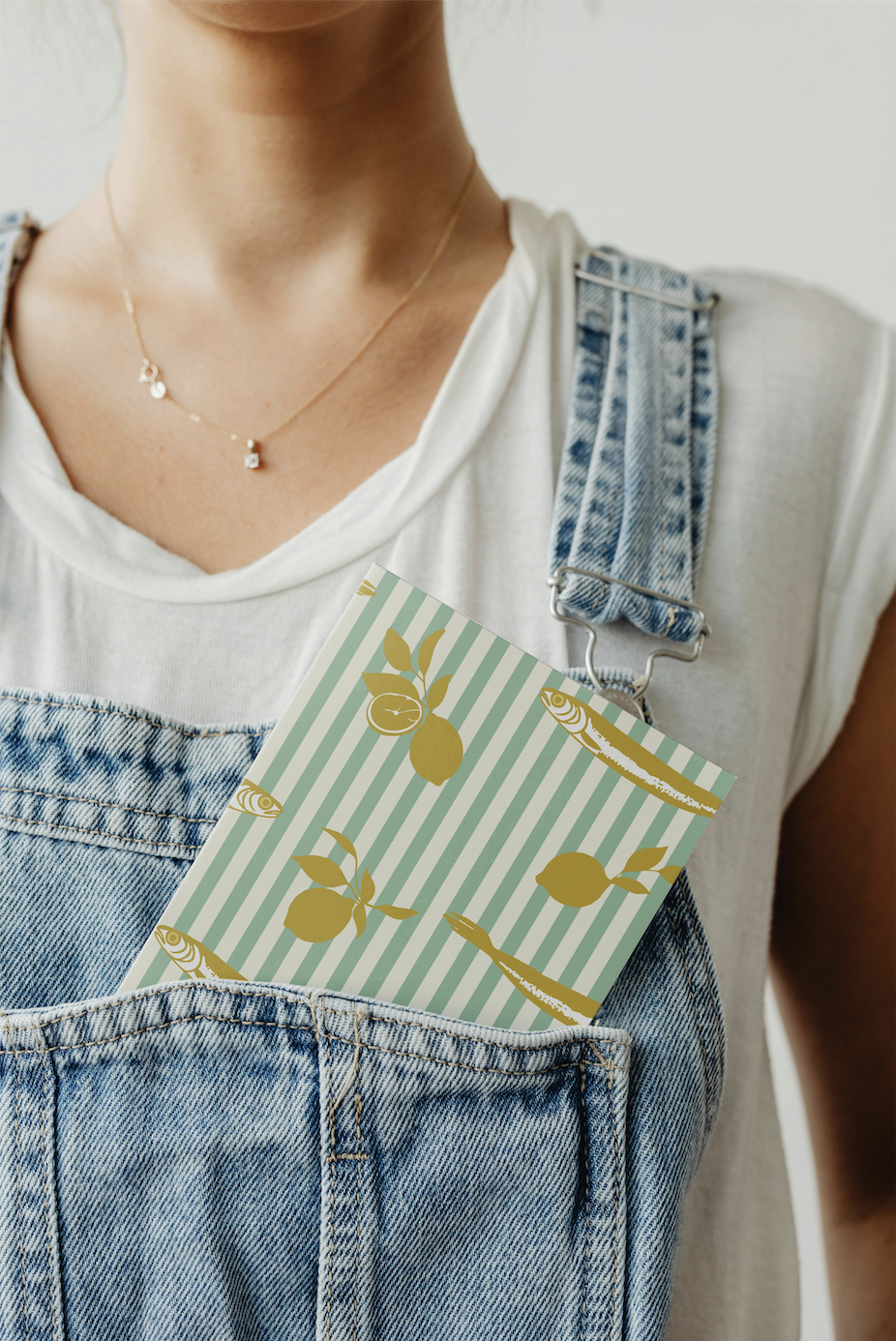 Close-up of a person wearing a white T-shirt, denim overalls, and a delicate gold necklace. A notebook with a lemon and fish pattern is tucked into the front pocket of the overalls.