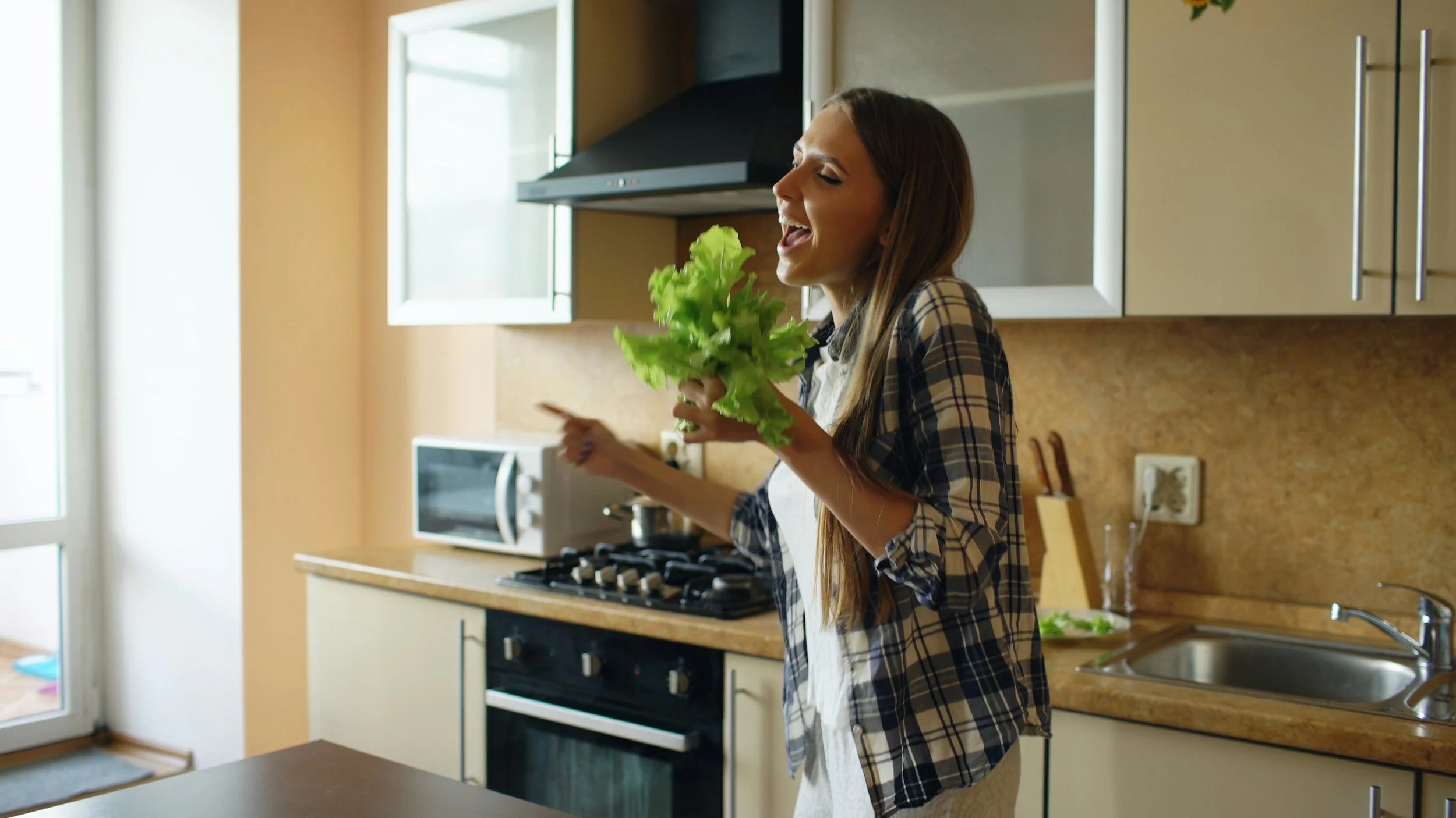 Woman holding a bunch of fresh lettuce in a kitchen with beige cabinets, a microwave, and a stove.
