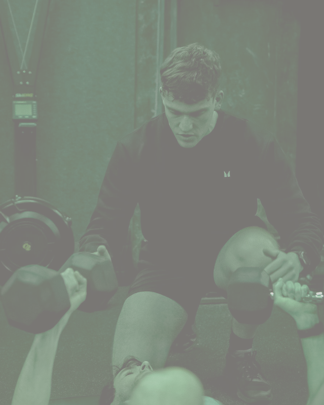 A fitness expert is assisting a client lying on the ground and lifting dumbbells during a workout session in a gym.