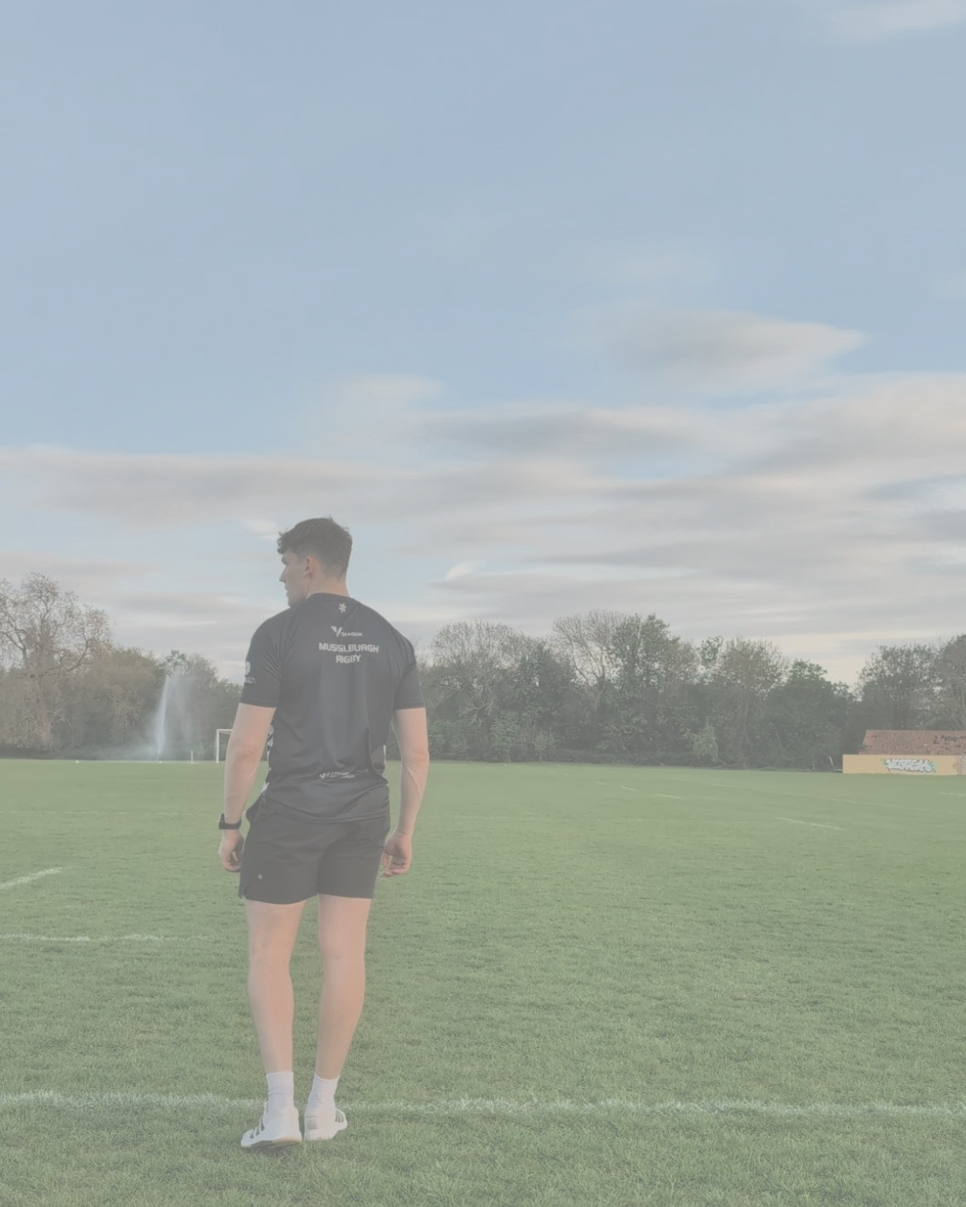 A rugby player wearing a black sports uniform stands on a grassy sports field with trees and a small building in the background. He is facing away from the camera under a partly cloudy sky.