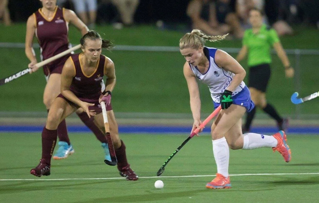 female playing international field hockey during a match, with one in a white and blue uniform controlling the ball and others in maroon uniforms trying to defend. strength and conditioning client