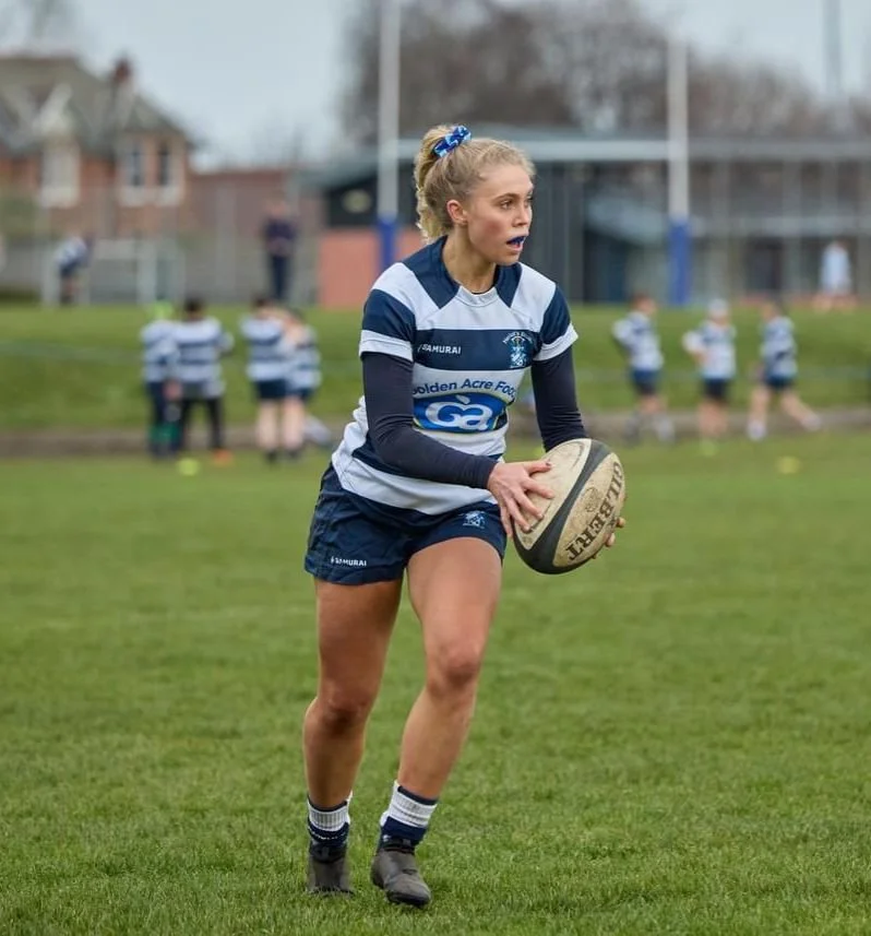 A semi-professional woman playing rugby on a grass field, holding a rugby ball, with rugby players in matching sports uniforms in the background. Physical Performance client