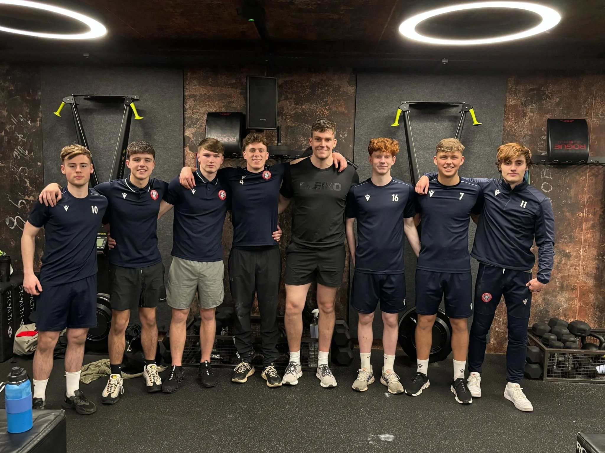 A group of seven young men and one adult man standing together in a gym, posing for a photo. The young men are in matching navy blue athletic shirts with numbers, while the adult man is wearing a black shirt. The background includes gym equipment and a textured wall.