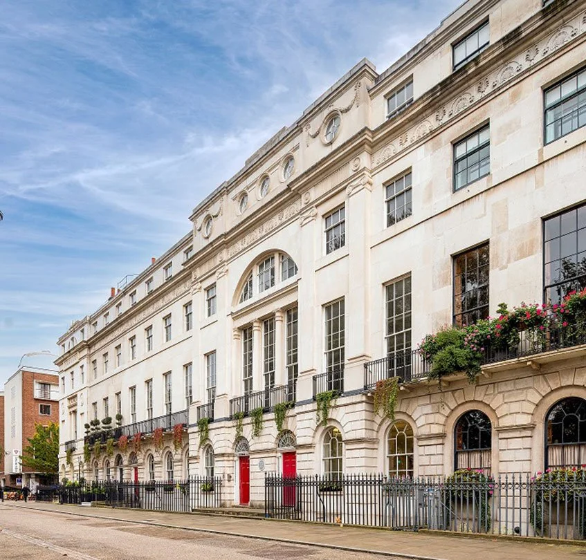 A historic white stone building with large arched windows, decorative architectural details, and black iron balconies with potted plants on a city street.