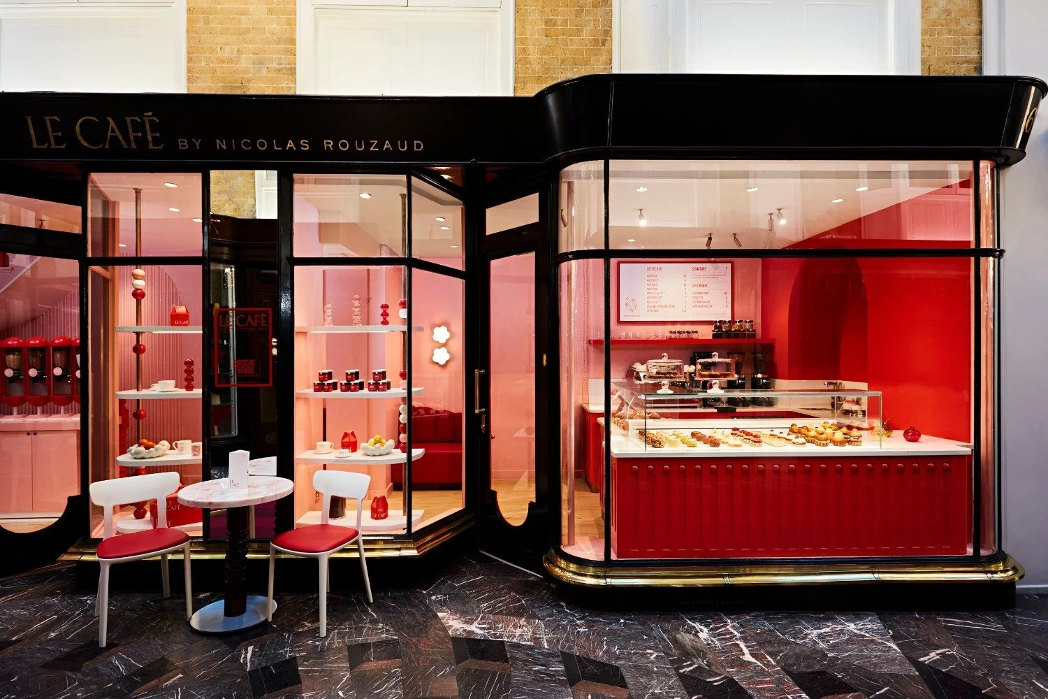 Exterior of a French bakery and coffee shop named Le Café by Nicolas Rouzaud with pink interior, display cases filled with pastries and desserts, and outdoor seating with two white chairs and a round marble table.