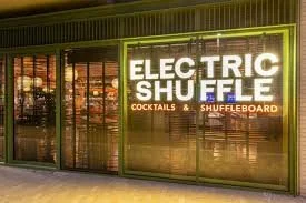 Storefront window with a sign reading 'Electric Shuffle' and smaller text for 'Cocktails' and 'Shuffleboard', illuminated at night.