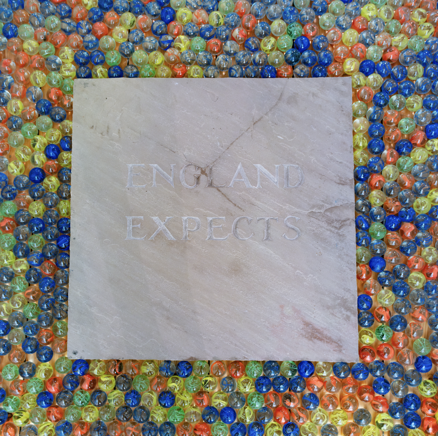 A square marble plaque with the words 'England Expects' engraved on it, surrounded by colorful marbles in shades of blue, green, yellow, and orange.