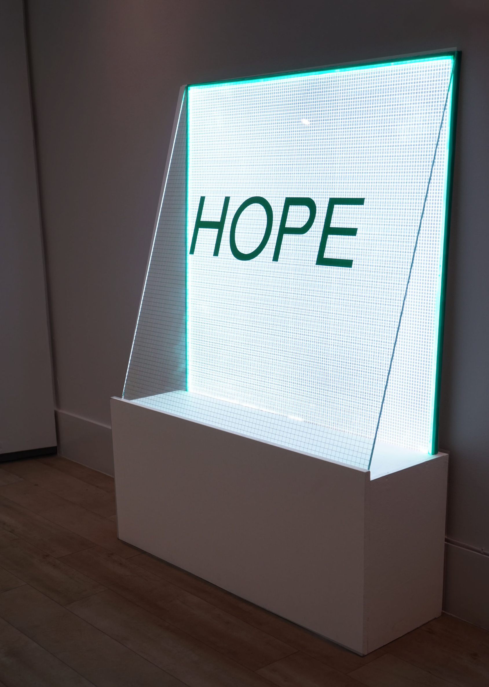 A white illuminated display with the word 'HOPE' written on it, placed on a wooden floor against a plain wall.