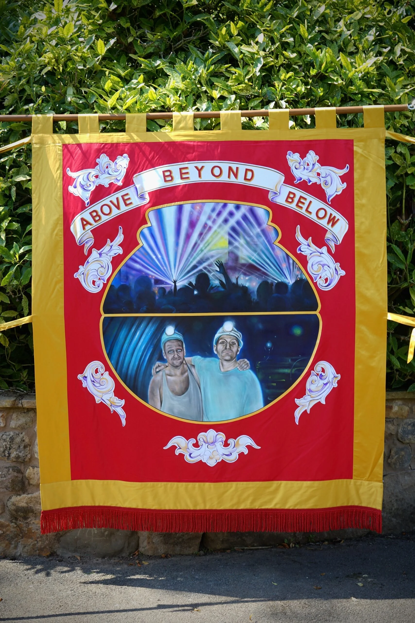 A colorful banner featuring a crowd at a concert or festival with vibrant laser lights in the background. The banner has a red and yellow border with decorative white scrollwork. The text reads "Above Beyond" on a ribbon at the top. Below, there is an illustrated scene of two people wearing headlamps, standing together, with one person resting their arm on the other's shoulder.