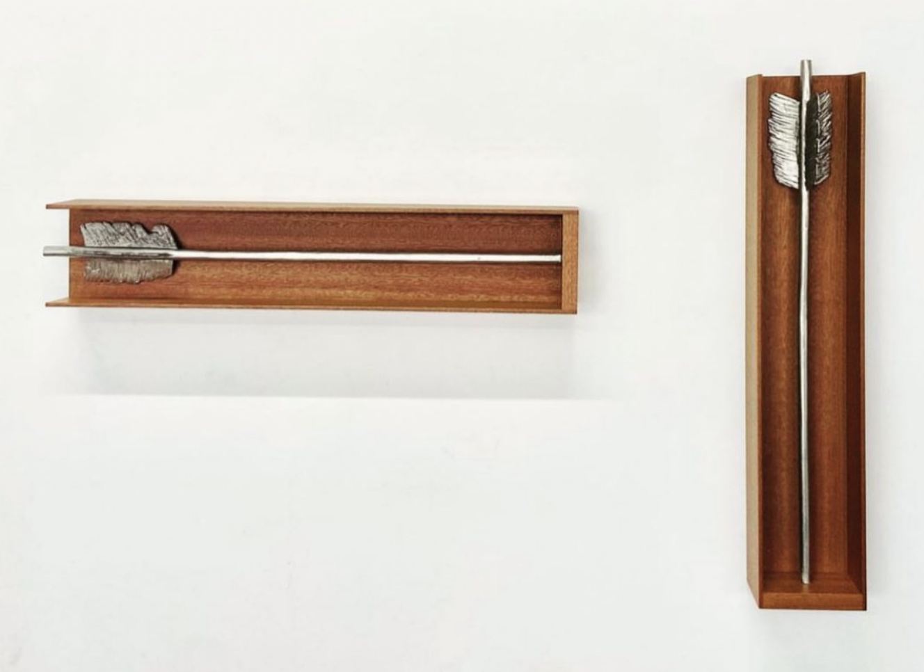 Two double-ended arrows, one on the wall and one in a wooden box, each with a feathered tail at each end.