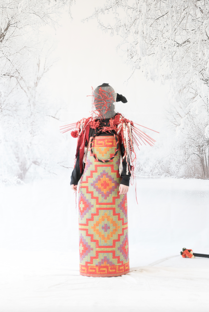 Person dressed in a colorful, woven traditional garment and elaborate mask, standing against a snowy winter backdrop with leafless trees.