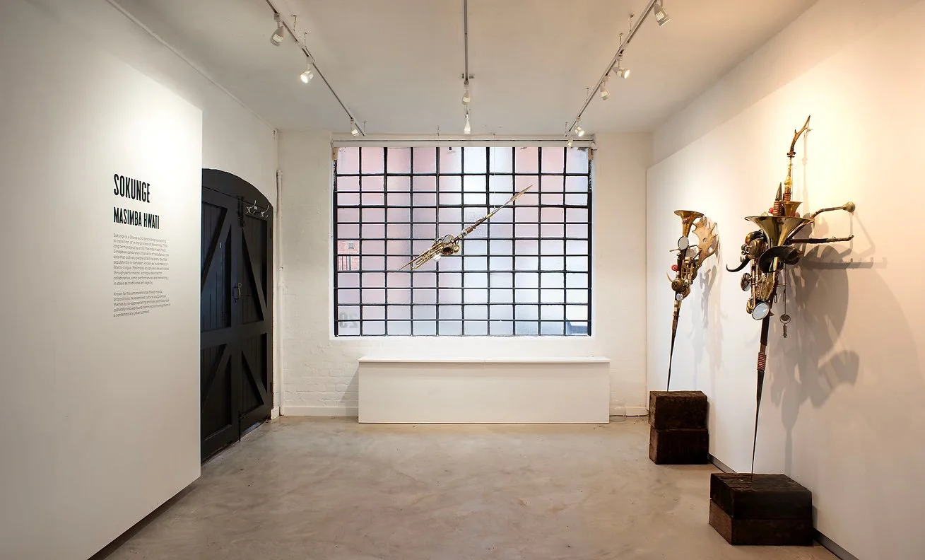An art gallery featuring sculptures on the right wall and a suspended sculpture in front of a large grid window, with white walls and a concrete floor.