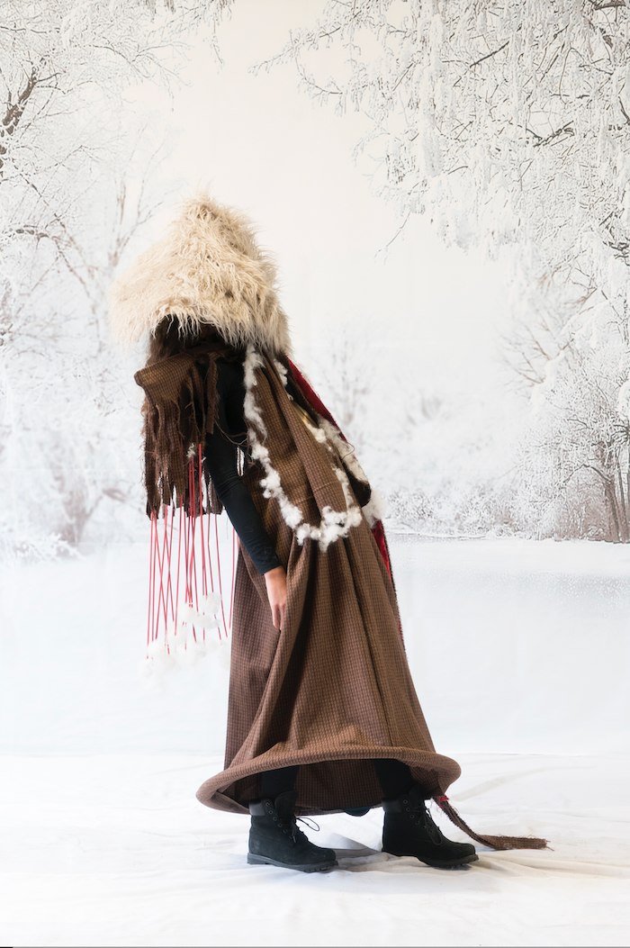 Person dressed in a winter costume with fur, draped in a brown patterned fabric, standing outdoors on snow with a frosted winter forest background.