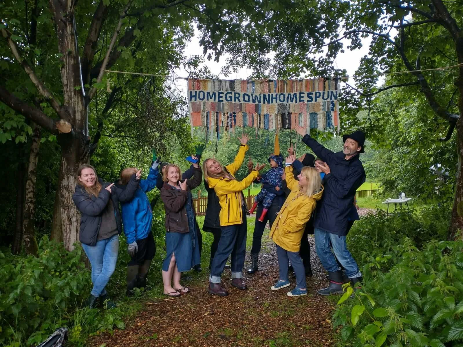 Group of people standing outdoors in front of a handmade sign that reads 'HOMEGROWN HOME SPUN', surrounded by green trees and foliage, celebrating outdoors. A project. with Patrick Grant