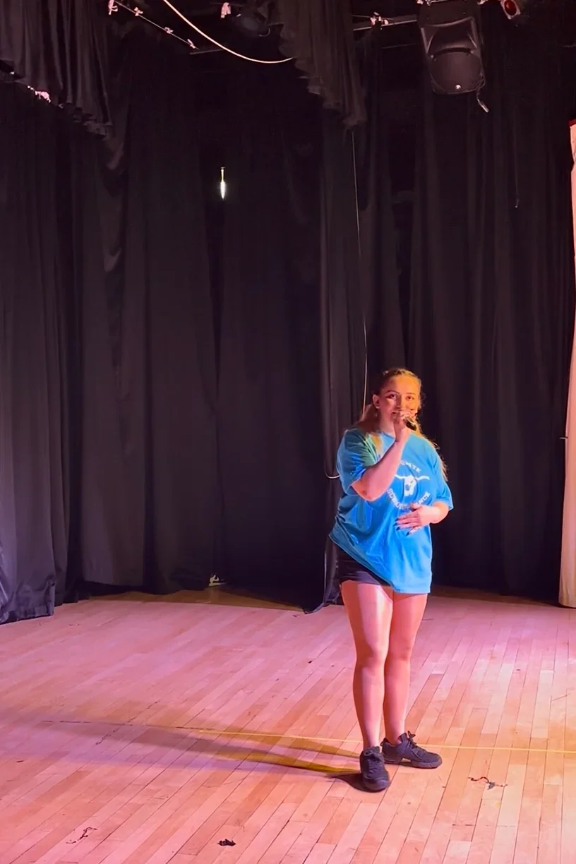 Miss Holly (school owner) singing on stage with a microphone, in front of black curtains, wearing a blue show t-shirt, black shorts, and sneakers.