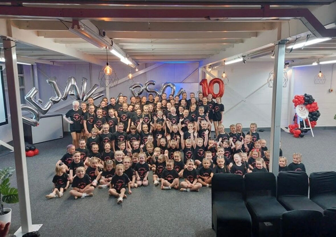 A large group of children and a few adults posing for a photo before a dance show in a room decorated with balloons that spell out 'DEJA VU 10'. The children are wearing matching uniform. There are balloon arrangements on the sides.