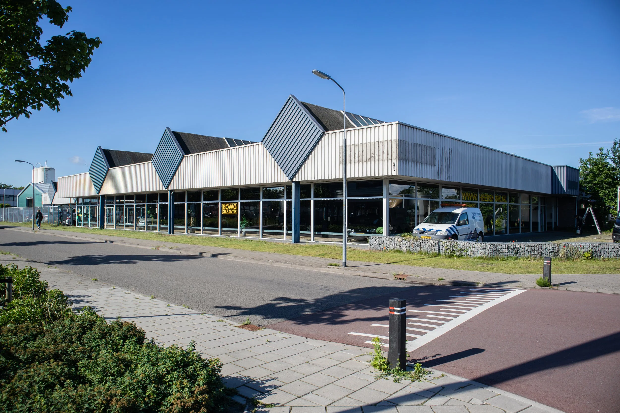 Winkelpand met grote ramen en witte metalen gevels, geparkeerde auto voor de winkel, straat met zebrapad, bomen en een heldere blauwe lucht.