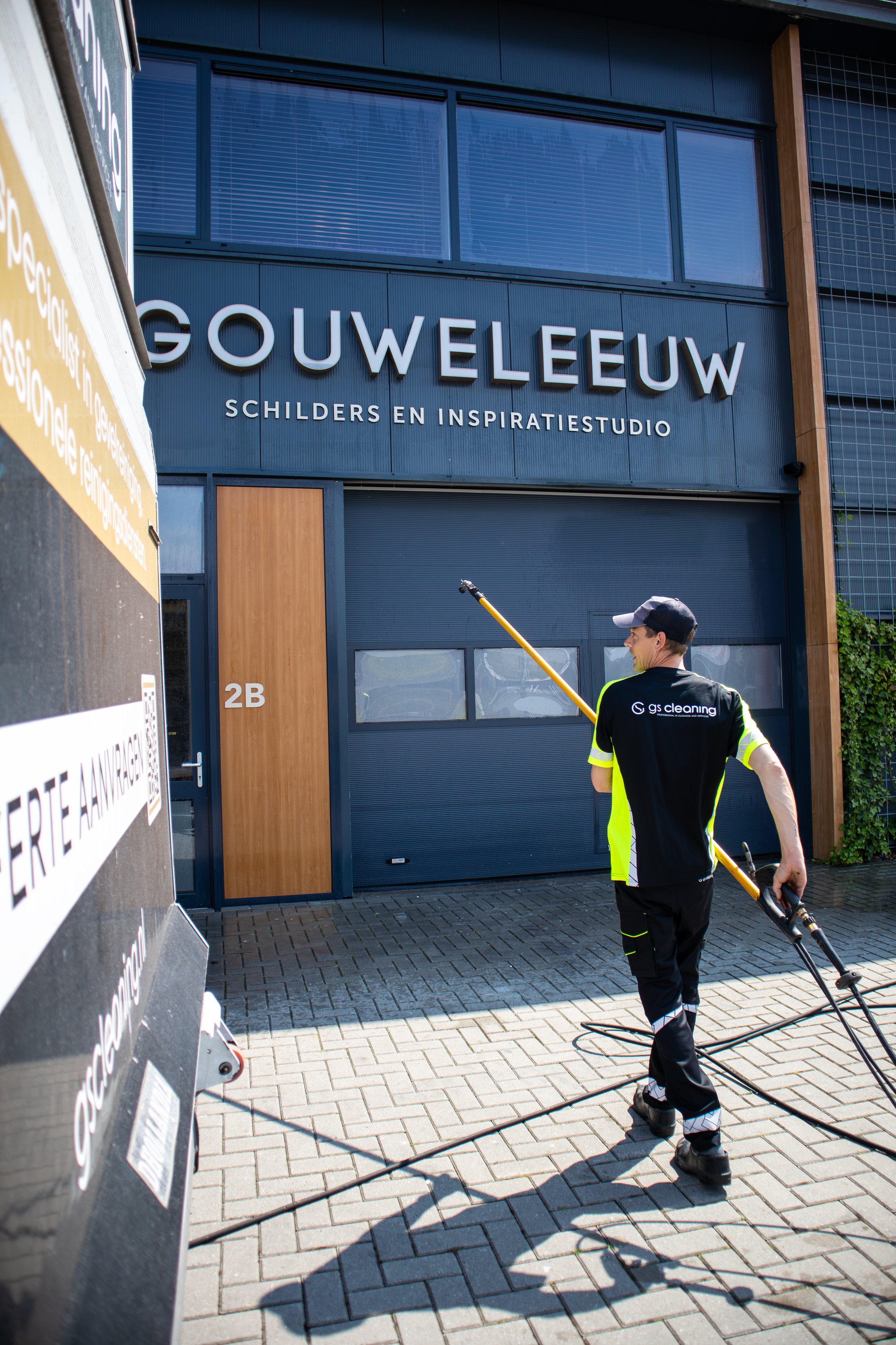 Man in werkkleding met gazonfrezen op de stoep voor een modern gebouw met blauwe gevel. Er staat een deur met het nummer 2B.