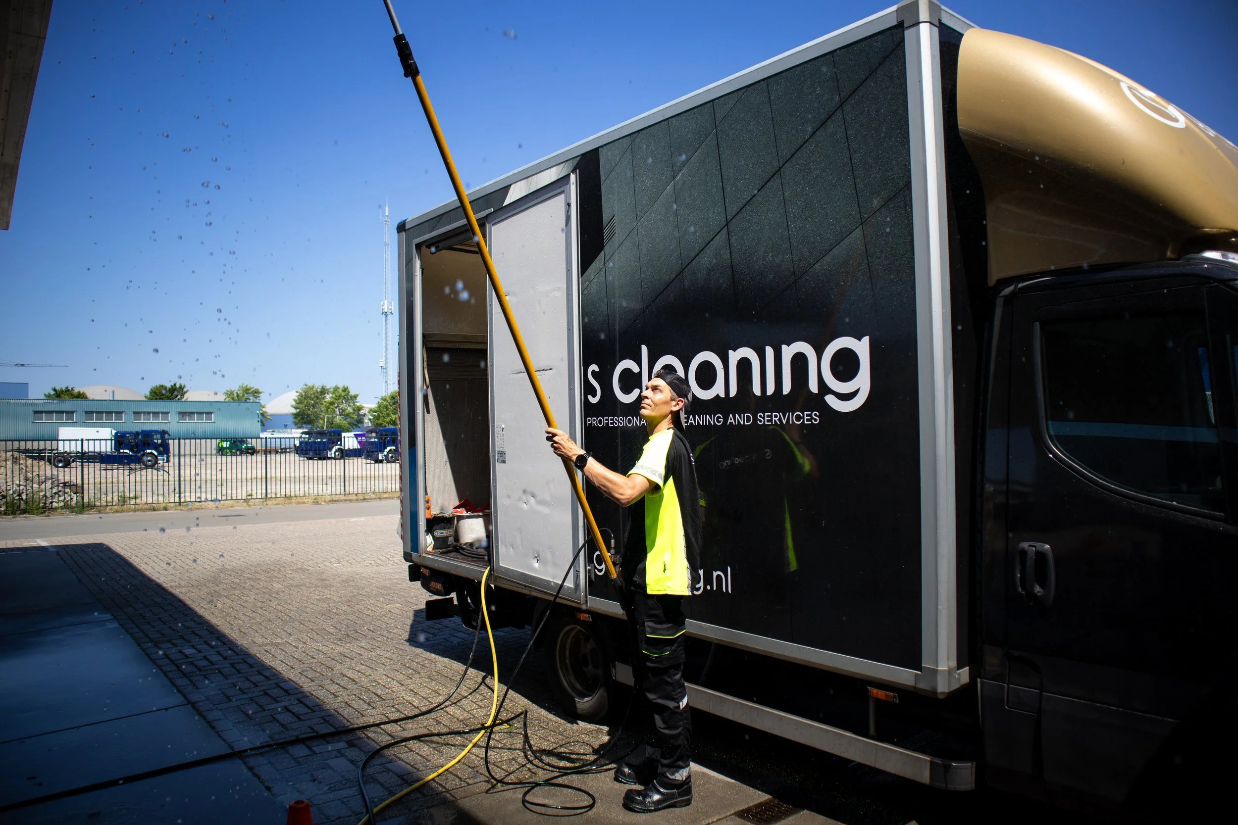 Een persoon in een gele veiligheidsjas en een zwart petje wast de buitenkant van een grote bus met een hogedrukspuit. De bus heeft het woord 's cleaning' en een webadres op de zijkant. Het is zonnig en er staan enkele auto's en gebouwen op de achtergrond.