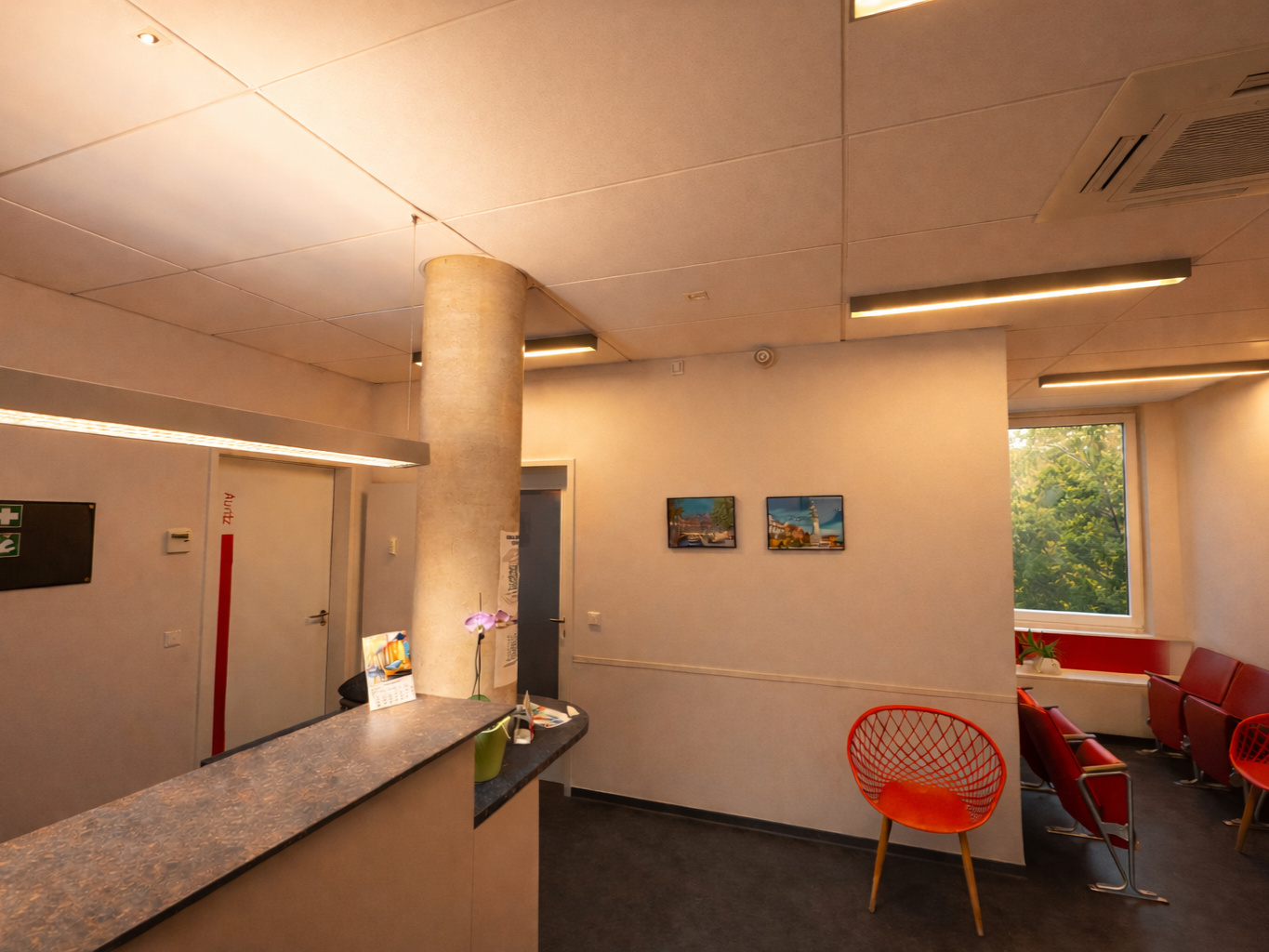 Innenansicht eines Wartezimmers mit roten Sesseln, einem Fenster mit Blick auf Bäume, einem Empfangstresen mit einer Vase und einem Kalender, Wandbilder und eine Deckenleuchte.