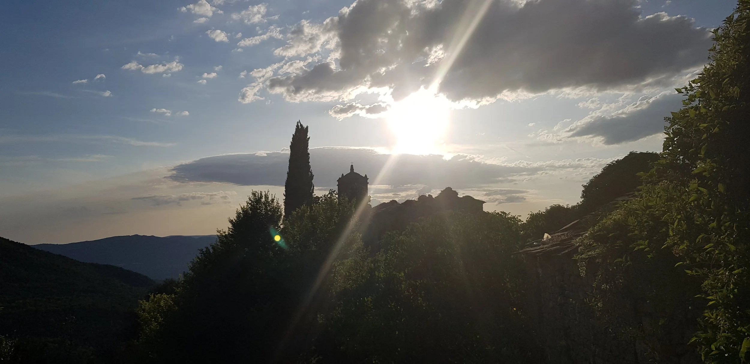 Paisaje con árboles y colinas, vista de una torre en la distancia, cielo con nubes y el sol brillando, reflejo de luz en el aire.