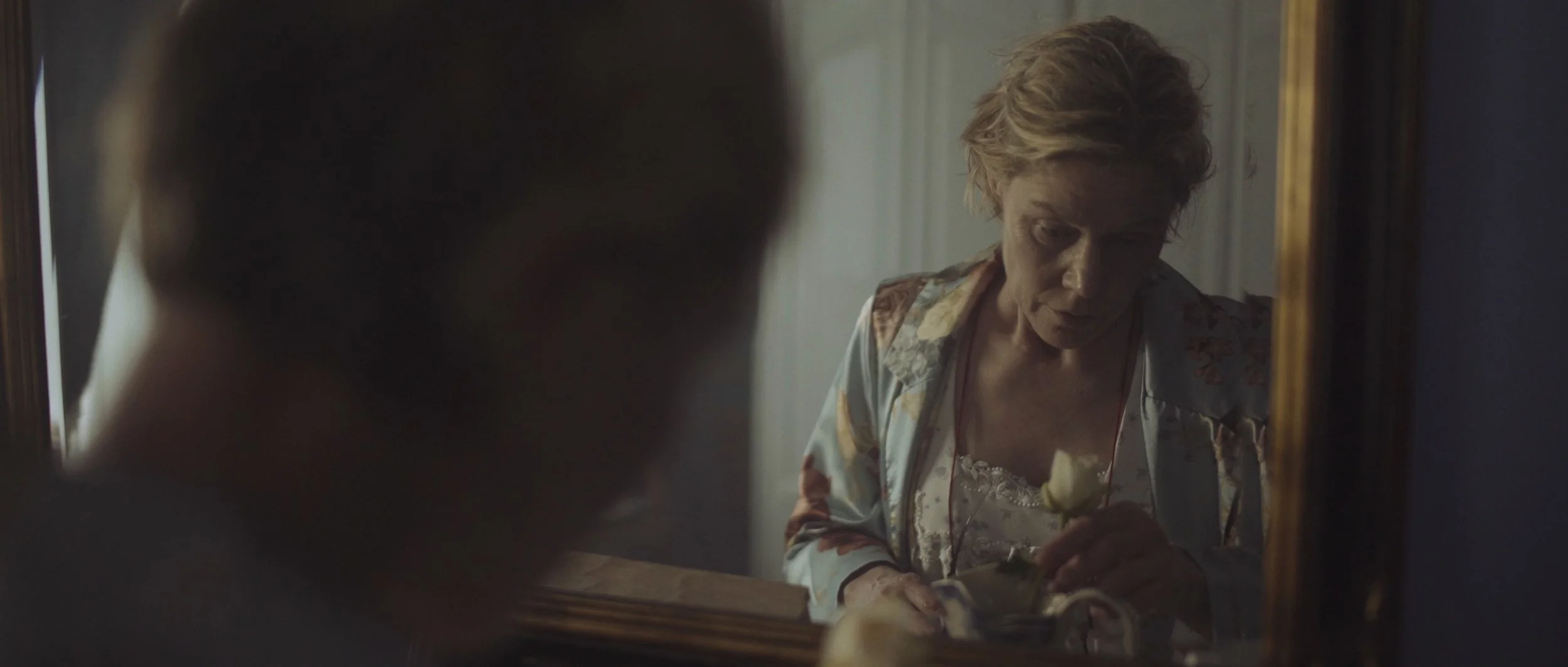 A woman looks at a flower as we see her reflection in the mirror