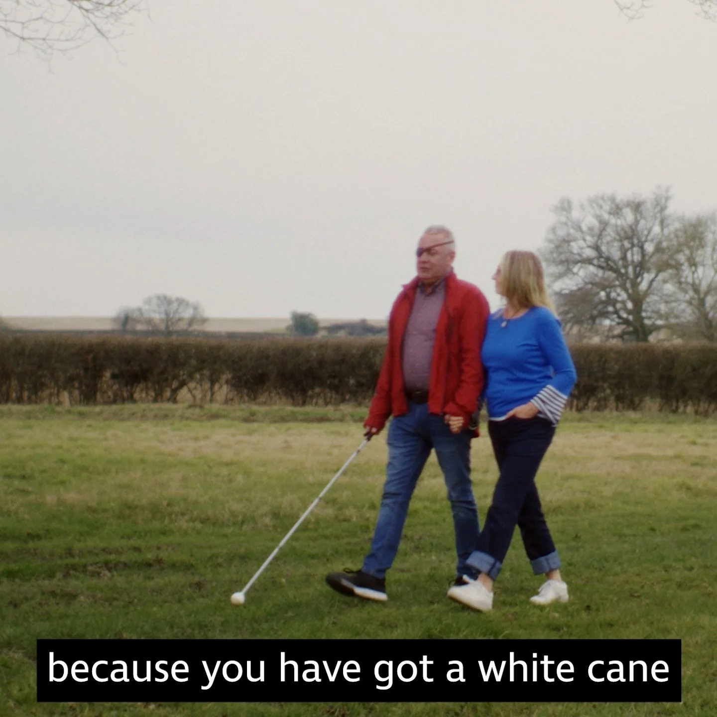 Pete and his wife walk in a field. Pete is holding a walking aid, RNIB