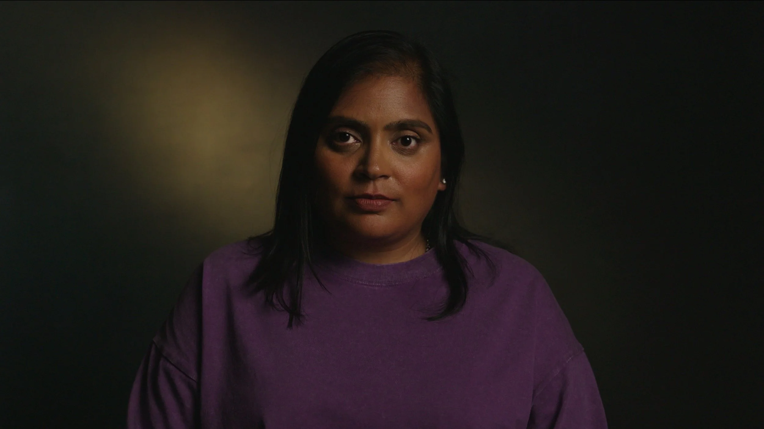 A woman is interviewed in a dark setting with moody lighting