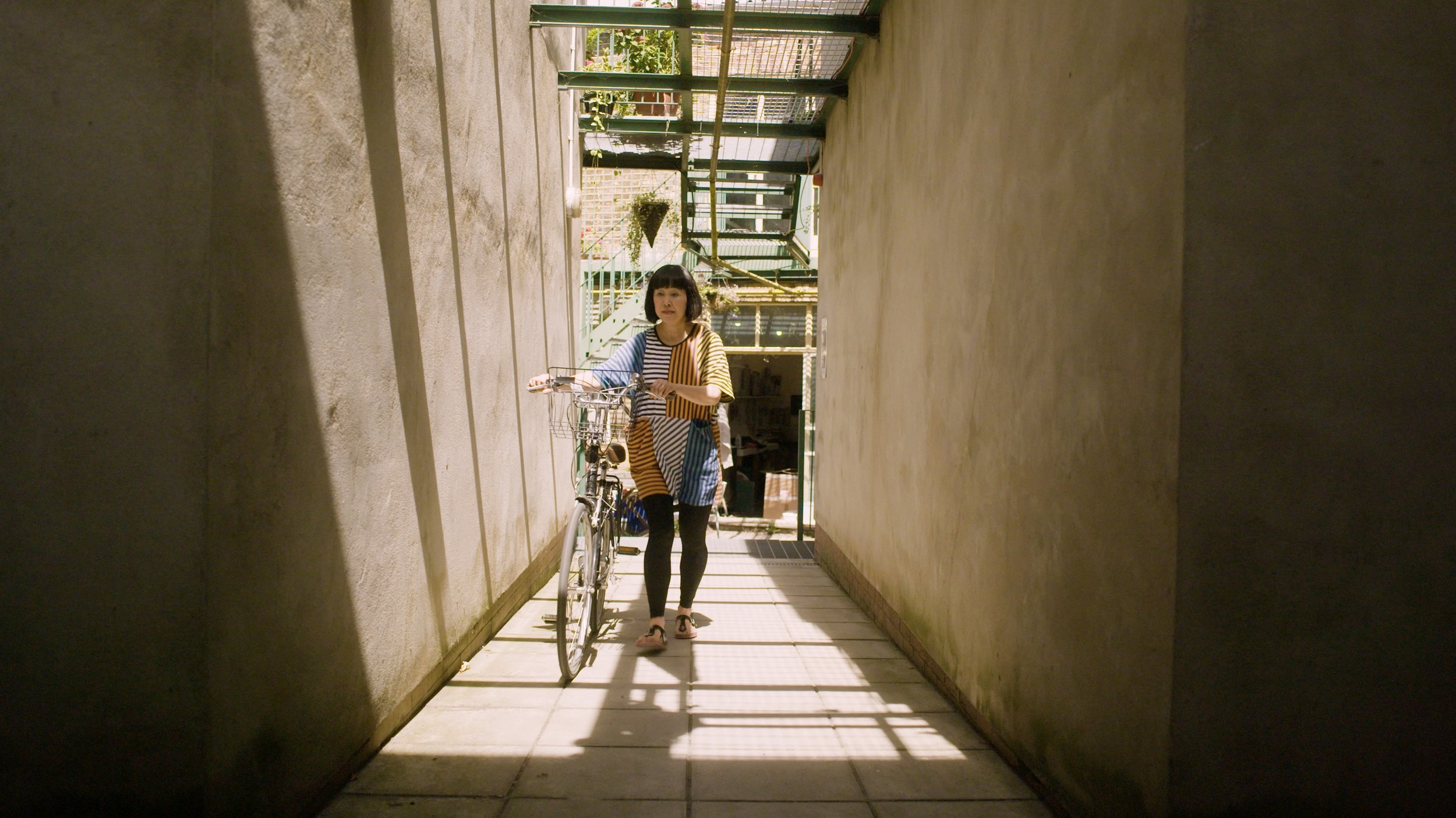 Ikuko Iwamoto walks with her bike down an outside corridor