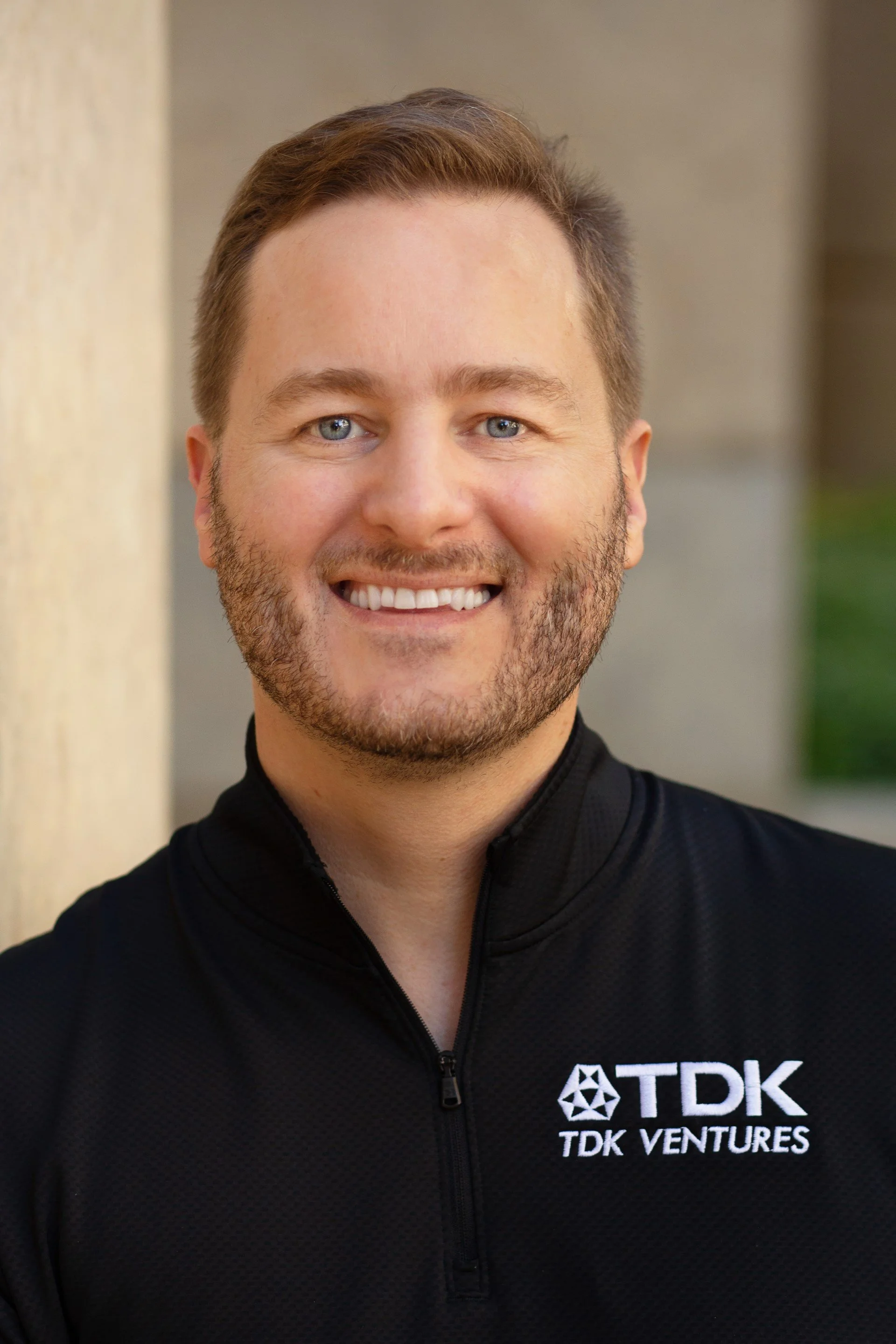 A smiling man with light brown hair and a short beard, wearing a black TDK Ventures jacket, standing outdoors with a blurred background.