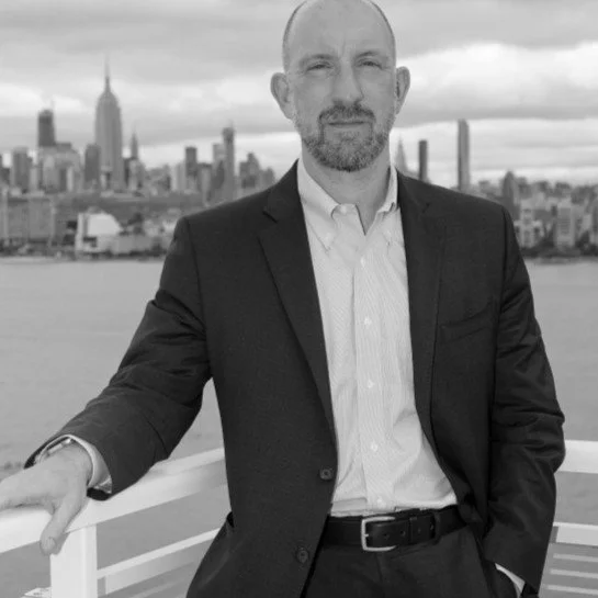 A man in a suit leaning on a railing with the New York City skyline in the background.