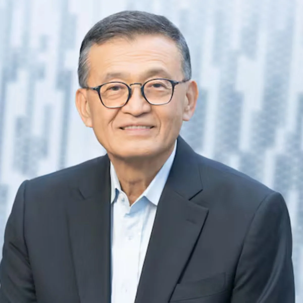 Portrait of an older man wearing glasses, a dark suit, and a light-colored collared shirt, smiling, with a light, textured background.