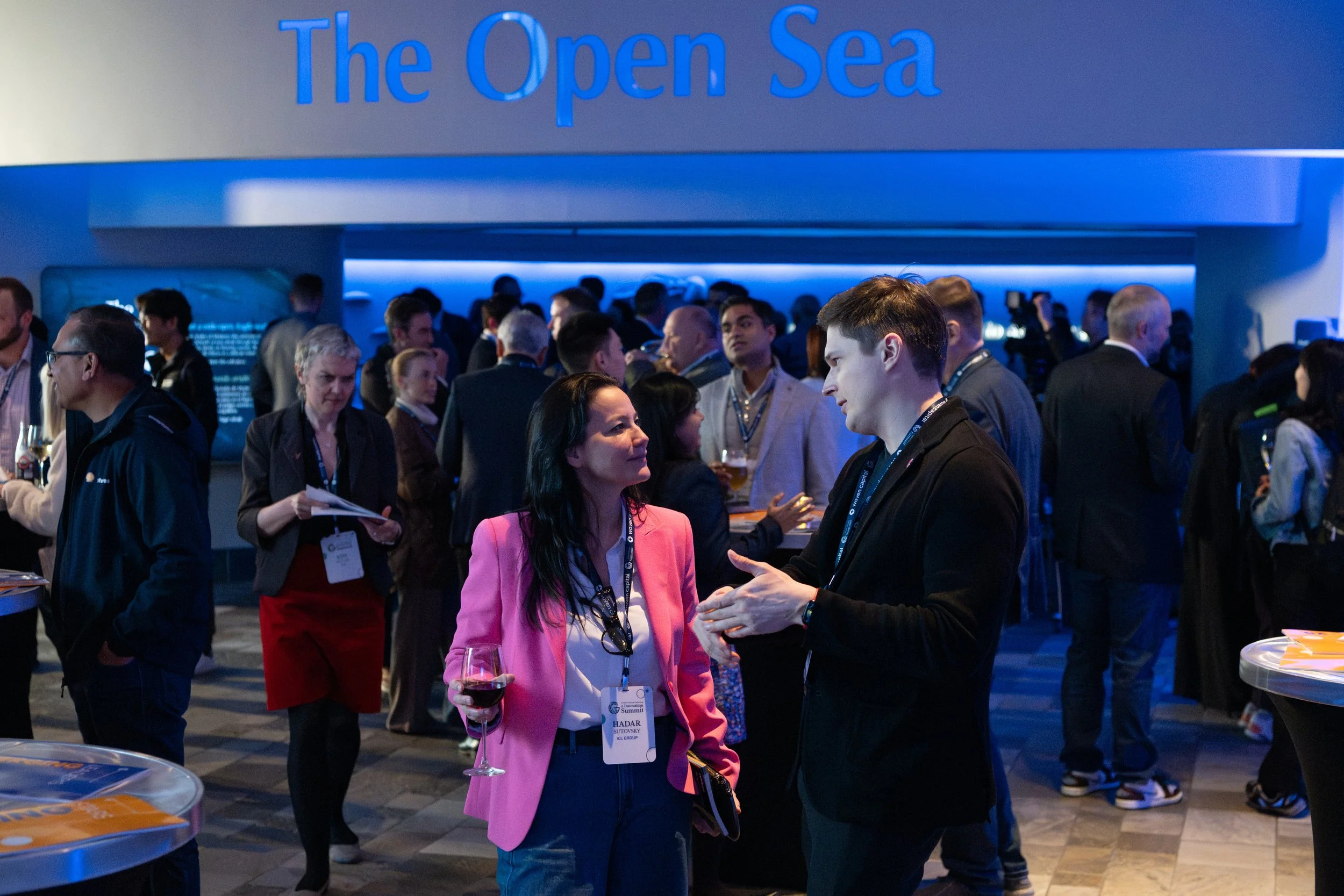 People attending a conference or event titled 'The Open Sea'. Two individuals in the foreground, a woman with long dark hair in a pink blazer holding a glass of wine talking to a young man, are engaged in conversation. The background shows more people mingling and networking in a dimly lit, modern venue with blue lighting.