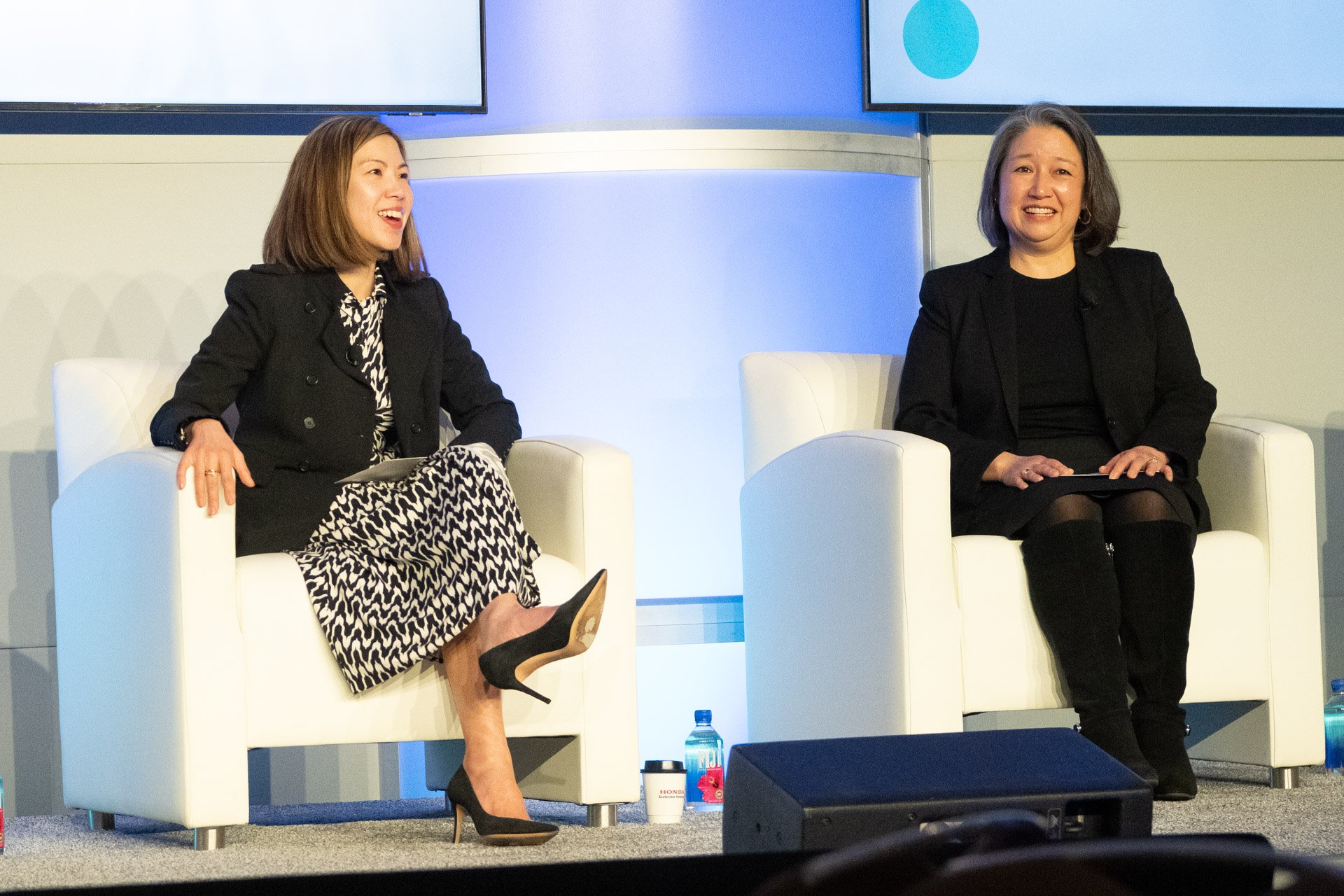 Two women sitting in white armchairs on a stage, engaging in conversation during a panel discussion or interview. There are large screens in the background displaying a blue circle on a white background.
