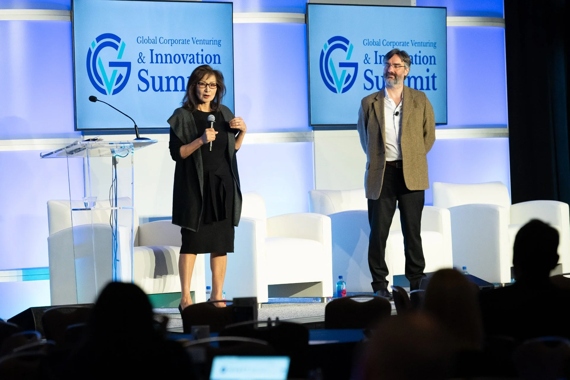 Two speakers, a woman and a man, stand on stage at the Global Corporate Venturing & Innovation Summit. The woman is speaking into a microphone, while the man stands with his hands behind his back. The background features two screens displaying the event's logo and name.