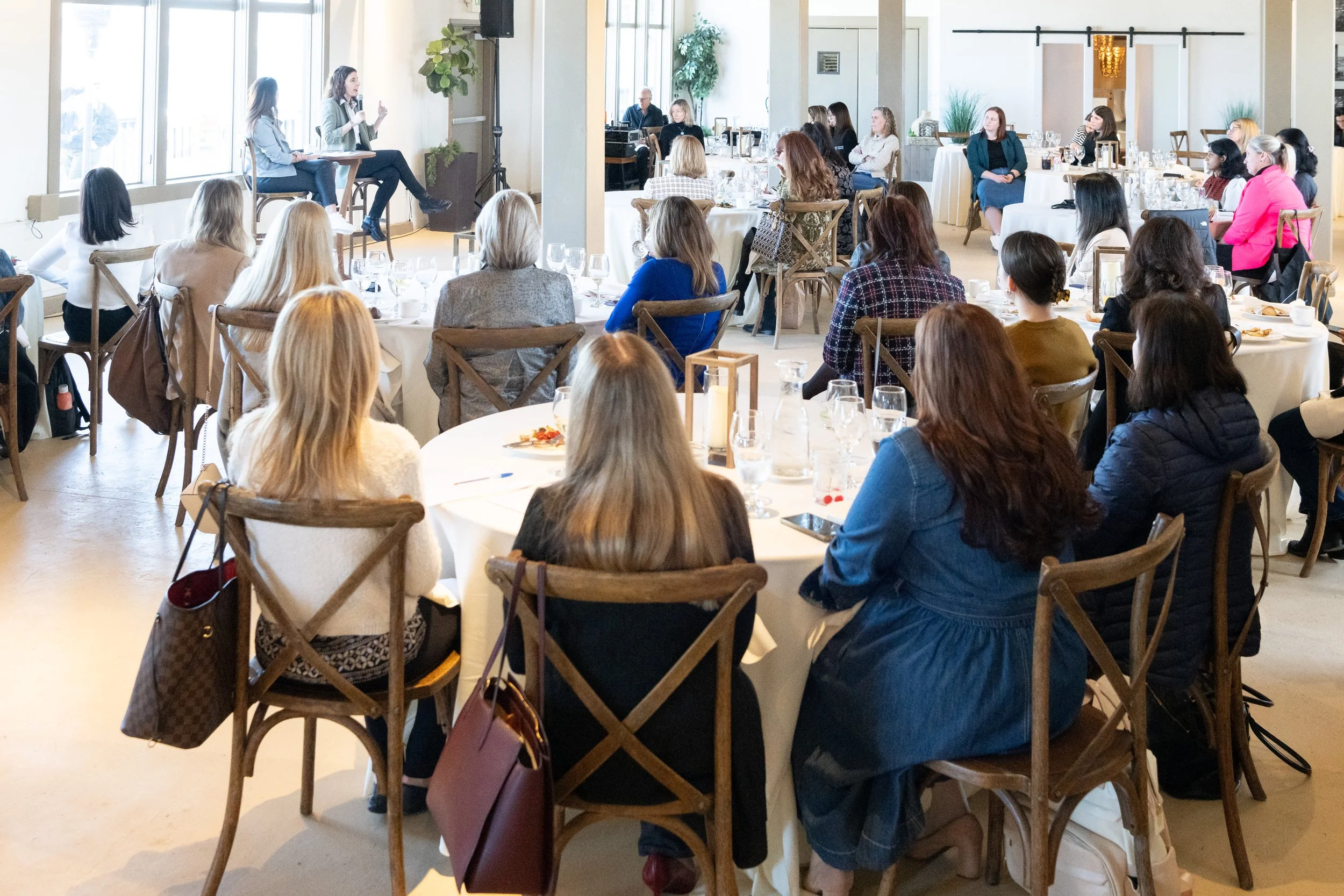 Women attending a conference or seminar in a bright, well-lit room with large windows, seated around round tables, listening to a woman speaker at the front.