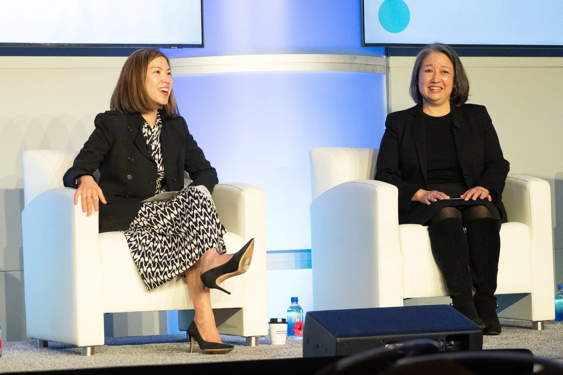 Two women sitting in white armchairs on a stage, engaging in conversation during a panel discussion or interview. There are large screens in the background displaying a blue circle on a white background.