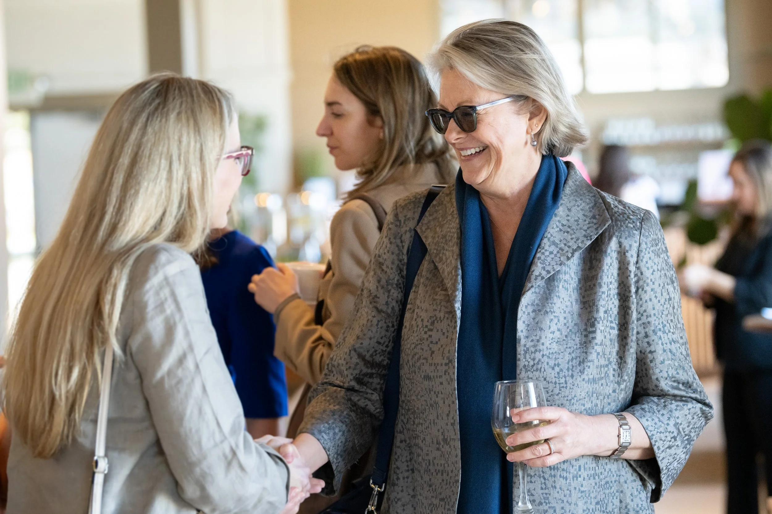 Two women shaking hands at a social gathering, one holding a glass of white wine, smiling, with others in the background.