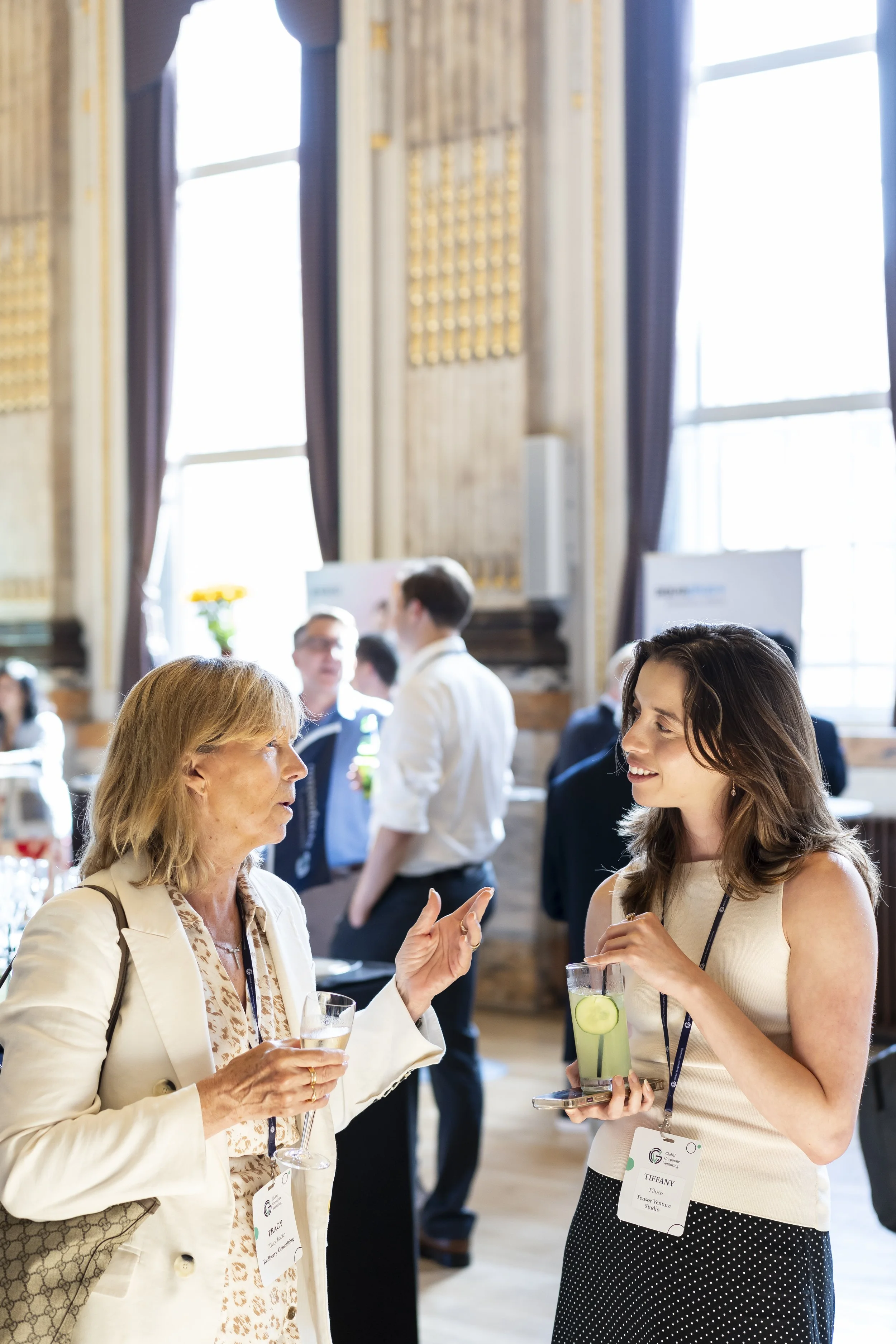 Two women with conference badges, one older with blond hair and one younger with brunette hair, talking at a social event. The older woman has a wine glass, and the younger woman has a drink with cucumber slices. They are in a bright room with large windows.
