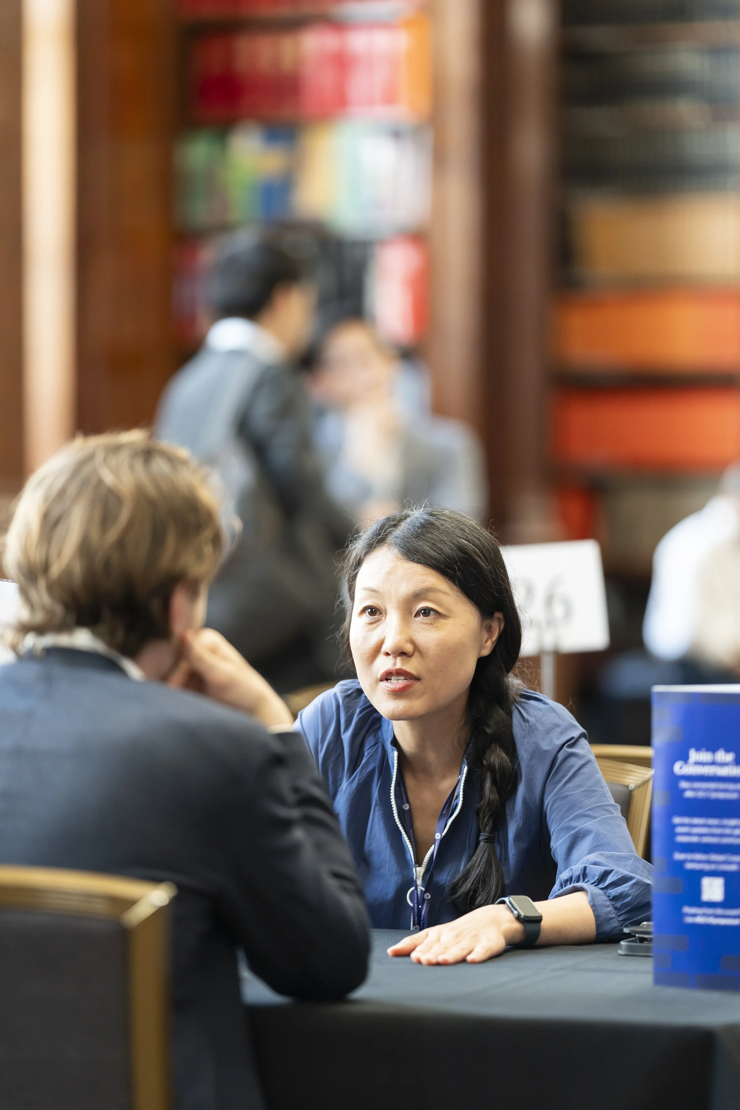 Two women engaged in conversation at a table in a library or bookstore, with bookshelves in the background