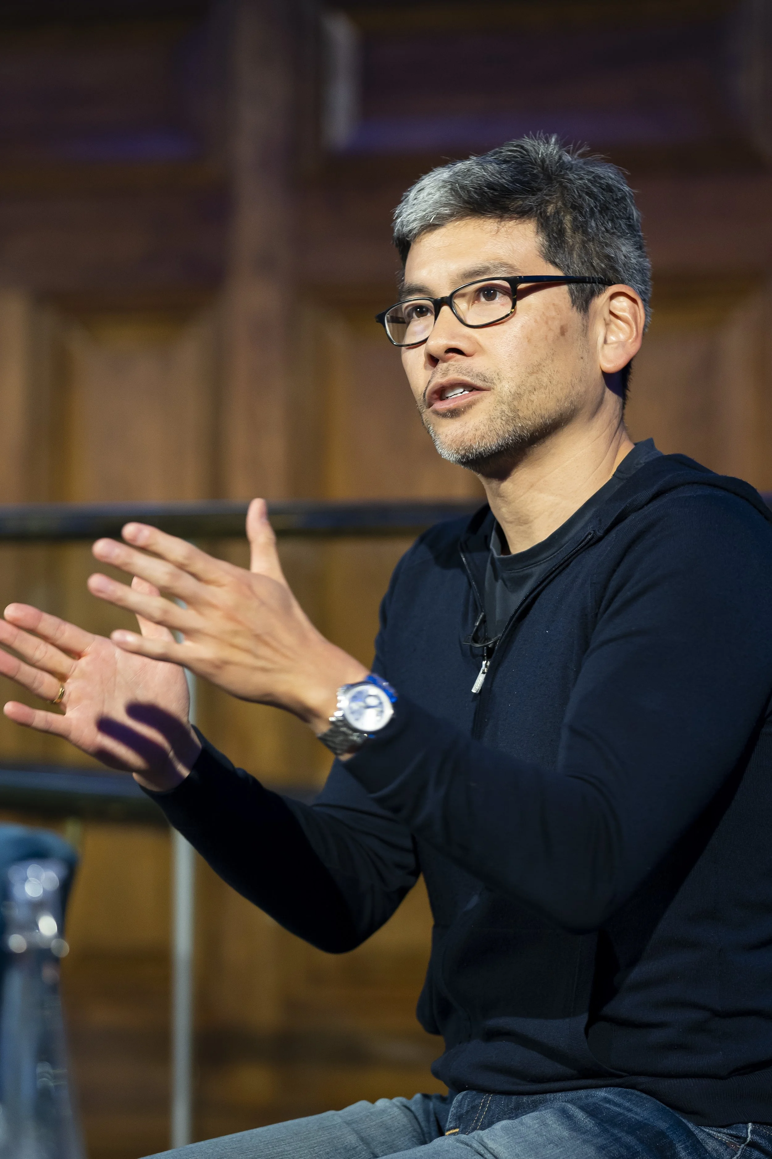 A man with glasses and graying hair is speaking and gesturing with his hands, wearing a black jacket and a watch, sitting in a room with wooden paneling in the background.